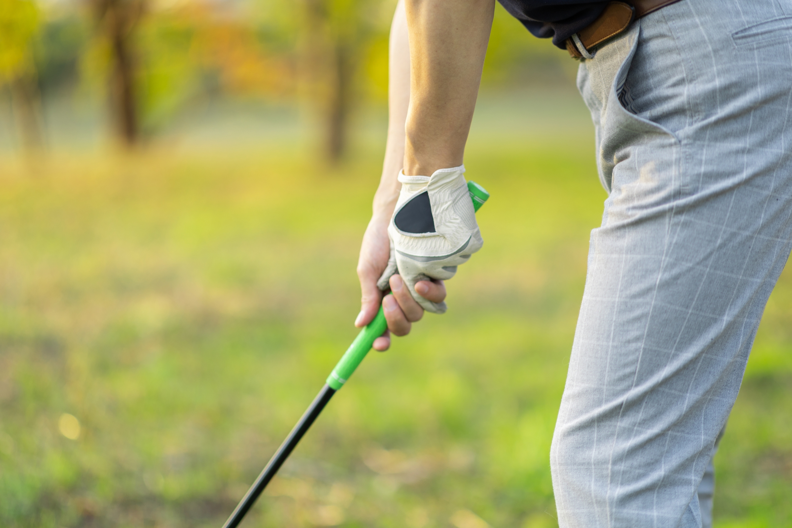 白いゴルフグローブをはめた手が緑のグリップのクラブを握り、グレーのスラックスをはいた人物が芝生の上で構えているクローズアップ。