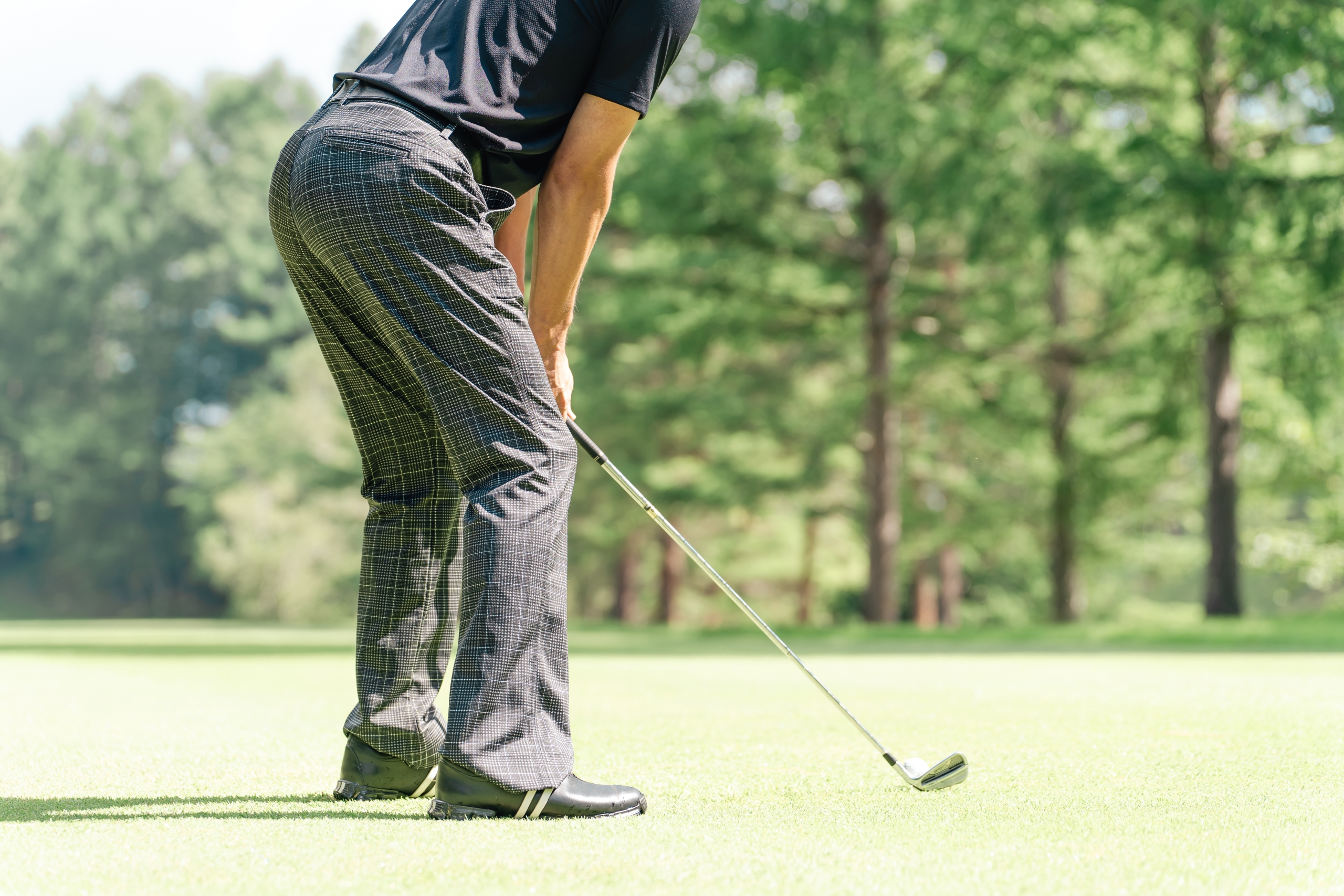 チェック柄のパンツと黒いシャツを着たゴルファーの下半身がグリーン上に立ち、アイアンでボールを打とうとしている木立のあるゴルフ場の風景。