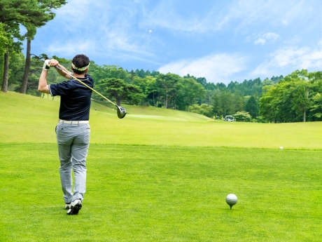 ティーグラウンドで黒いシャツとグレーのパンツを着たゴルファーがドライバーでスイングしてボールを打ち上げ、手前にティーに乗ったボールと奥に広がるフェアウェイと木立が見える風景。