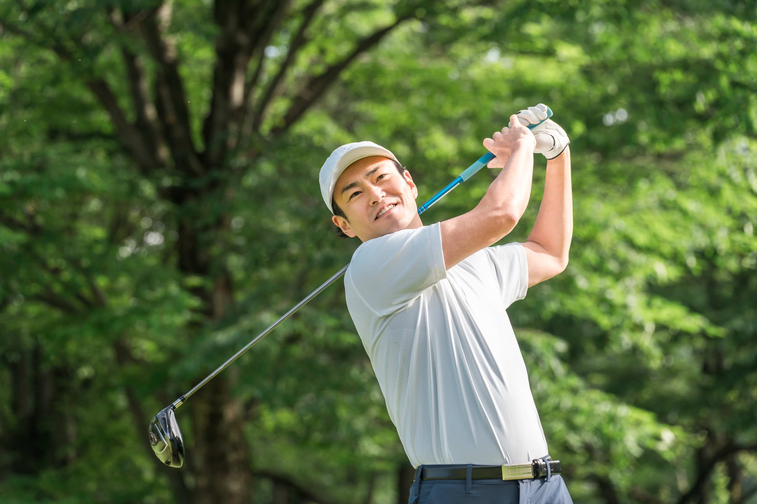 白い帽子とシャツ、グローブを着けた男性がドライバーをフルスイングして微笑む、緑の木々を背にしたゴルフ場の一場面。