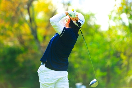 白い帽子とネイビーのポロシャツ、白いパンツを着たゴルファーがドライバーでフルスイングしている、背景は緑の木々がぼかされた屋外のコース。