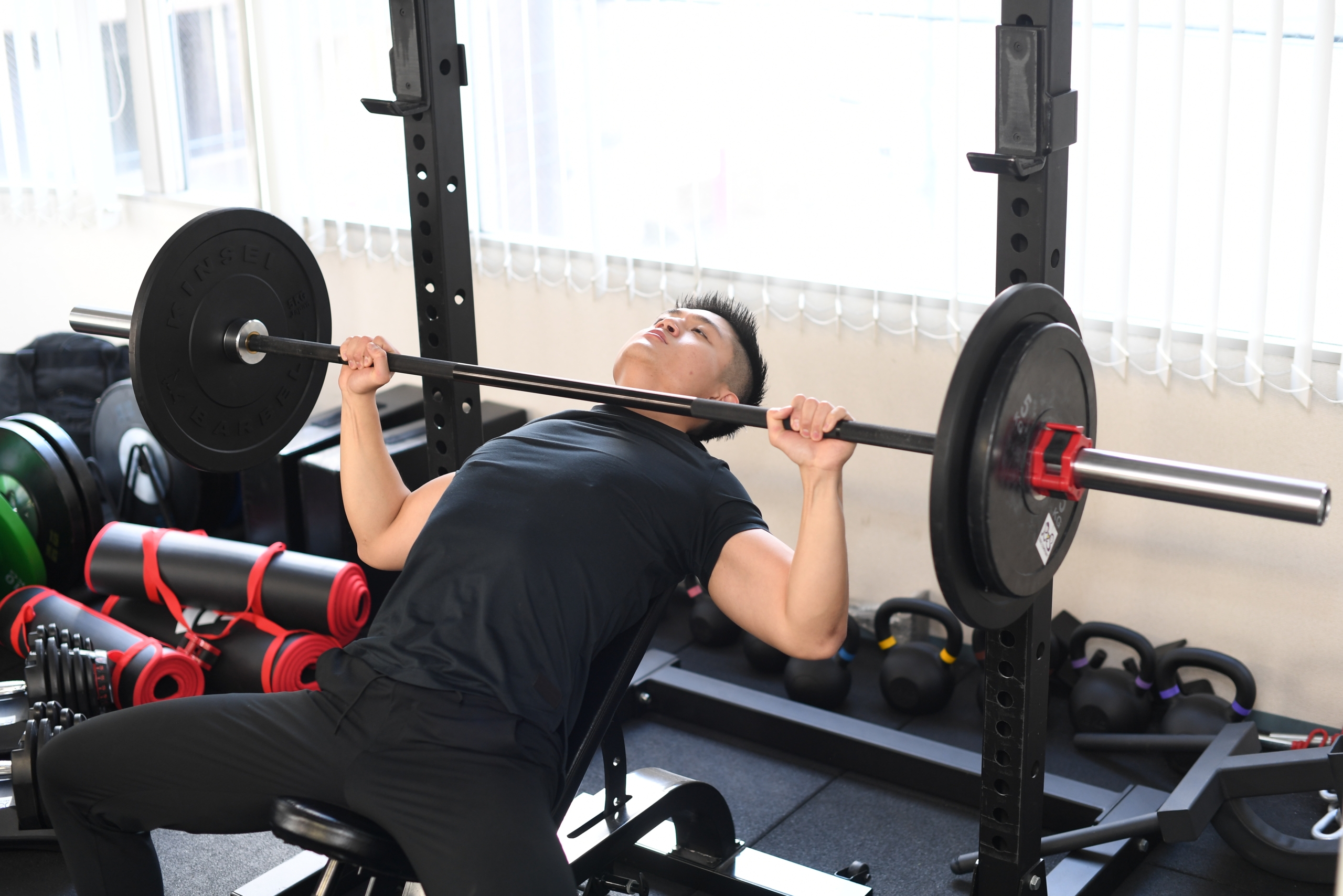 インクラインベンチに仰向けになりバーベルを持ち上げてベンチプレスをする男性、左右に重りの付いたバーと背景にケトルベルやヨガマットなどの器具が並ぶジムの室内。