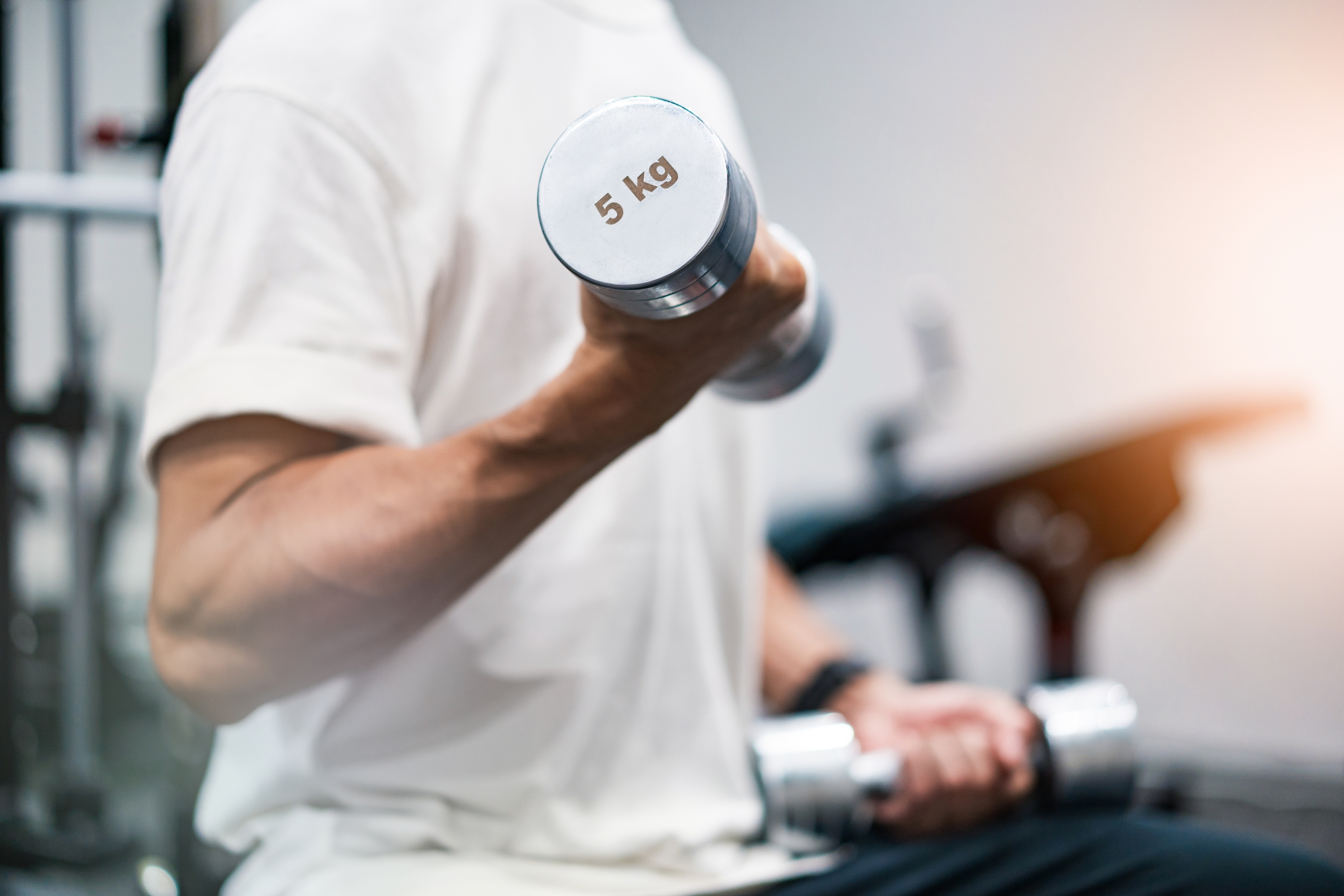 ジムで白いTシャツを着た男性が、5kgのダンベルを両手に持って筋力トレーニングをしている様子。