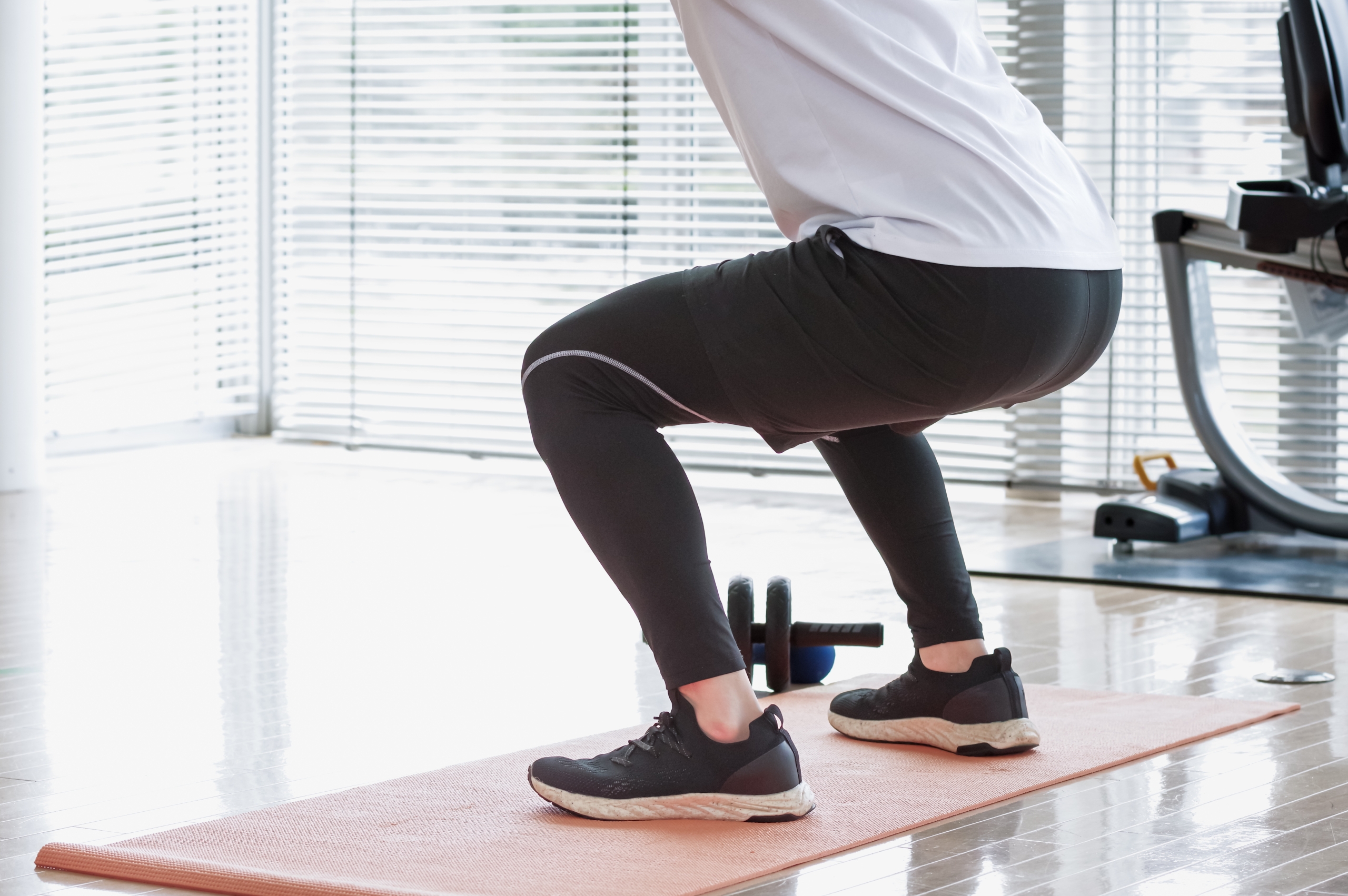 ジムで、トレーニングウェアを着た人がヨガマットの上でスクワットをしている様子。